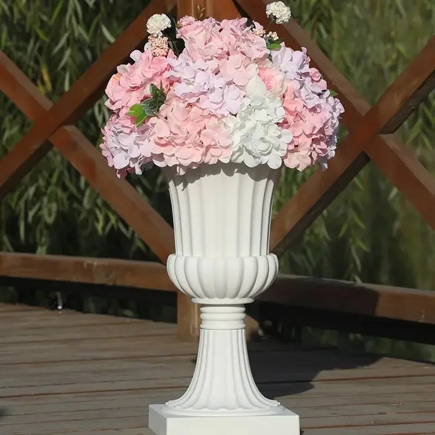Vase médicis blanc avec des hortensias roses photographié en extérieur entouré de végétation