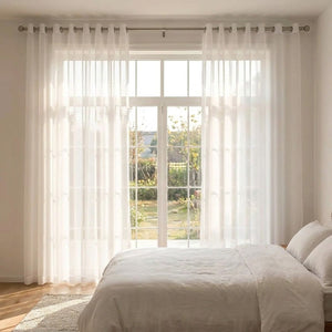 Voilage blanc encadrant une baie vitrée dans une chambre lumineuse avec lit blanc