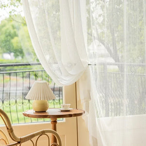 Voilage tulle blanc suspendu près d'une table en bois avec lampe et vue sur la verdure